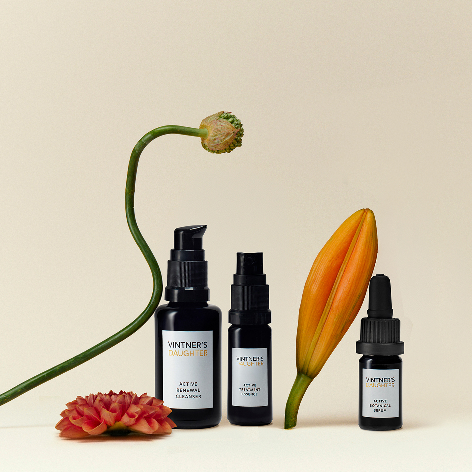 three black Vintner's Daughter skincare bottles arranged on a warm cream background with decorative flowers: a long green stem with a bud, a red-orange flower, and an orange lily. The bottles include a cleanser, toner, and serum.