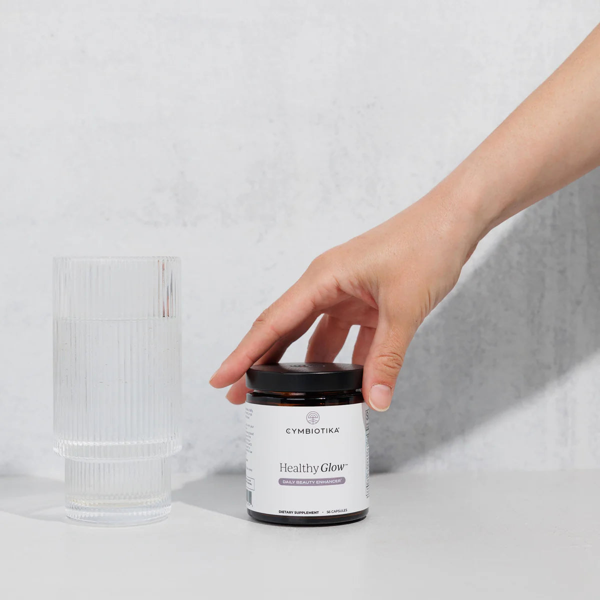 Hand holding a Cymbiota 'Healthy Glow' supplement jar on a light gray background