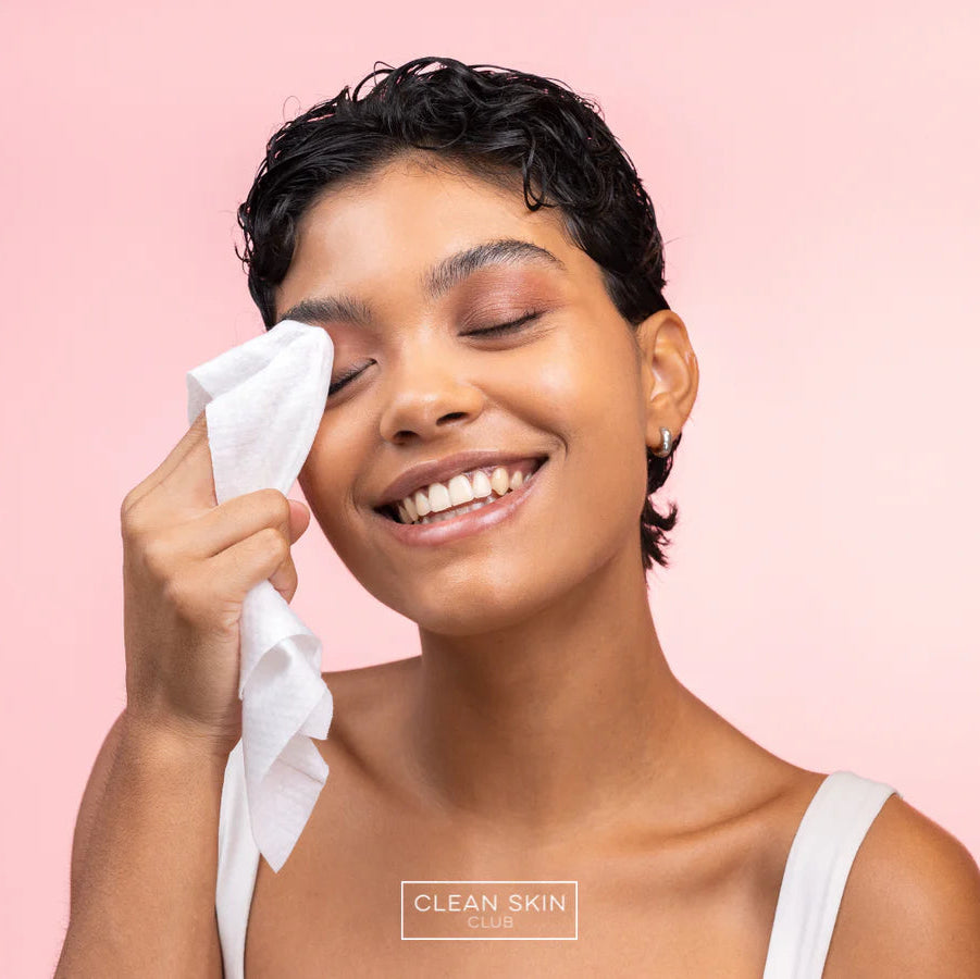 Woman cleaning her eye with a white cloth against a pink background, with 'Clean Skin Club' branding.
