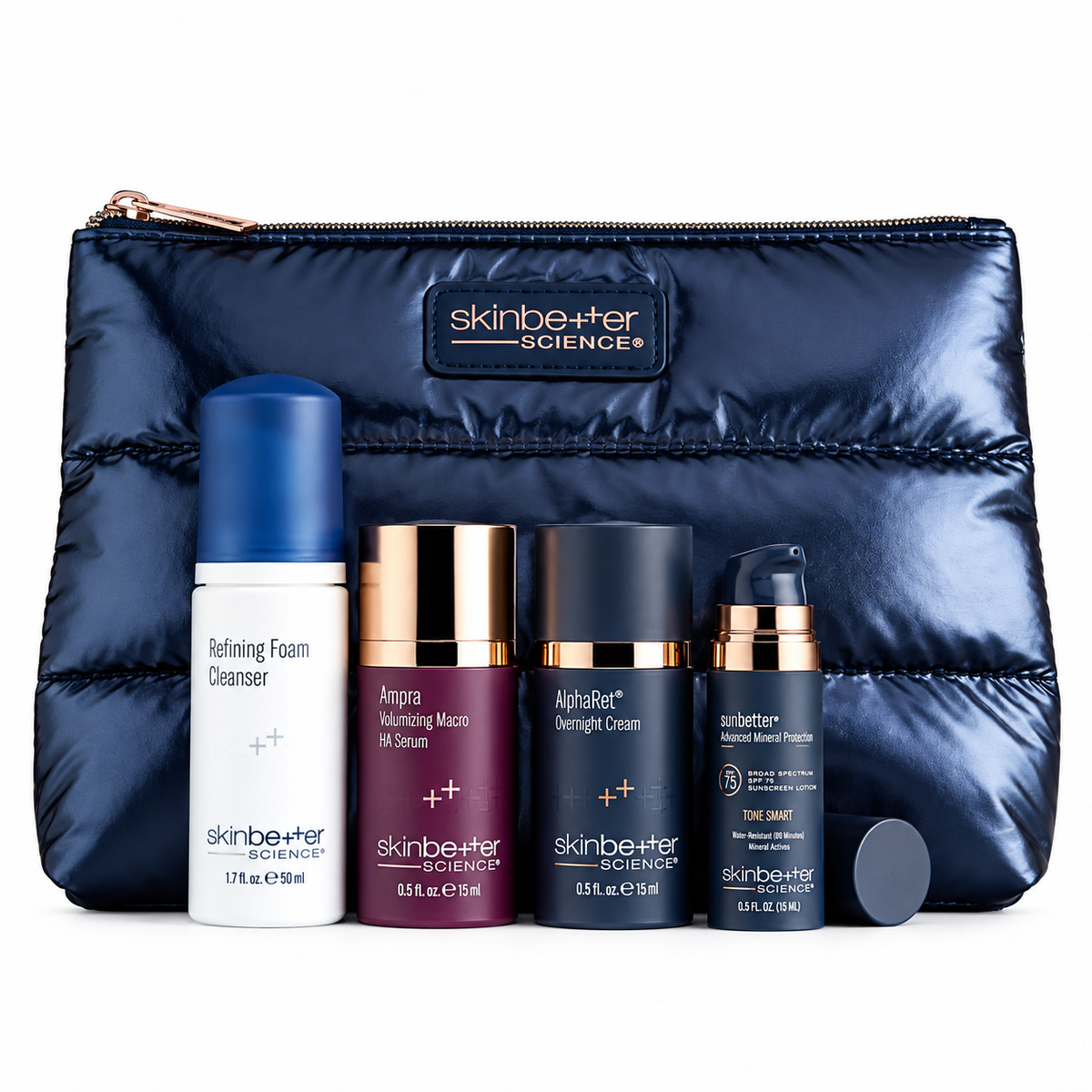A skincare gift set from Skinbetter Science featuring five products arranged in front of a navy blue puffer pouch. The set includes a white and blue Refining Foam Cleanser, a burgundy and gold Aspara Radiating Macro Oil Serum, a black and gold AlphaBet Overnight Cream, a black and gold Toner, and a black and gold Total Cream, with a small black cap lying in front of the pouch.