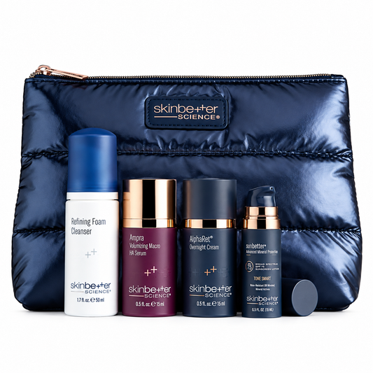 A skincare gift set from Skinbetter Science featuring five products arranged in front of a navy blue puffer pouch. The set includes a white and blue Refining Foam Cleanser, a burgundy and gold Aspara Radiating Macro Oil Serum, a black and gold AlphaBet Overnight Cream, a black and gold Toner, and a black and gold Total Cream, with a small black cap lying in front of the pouch.