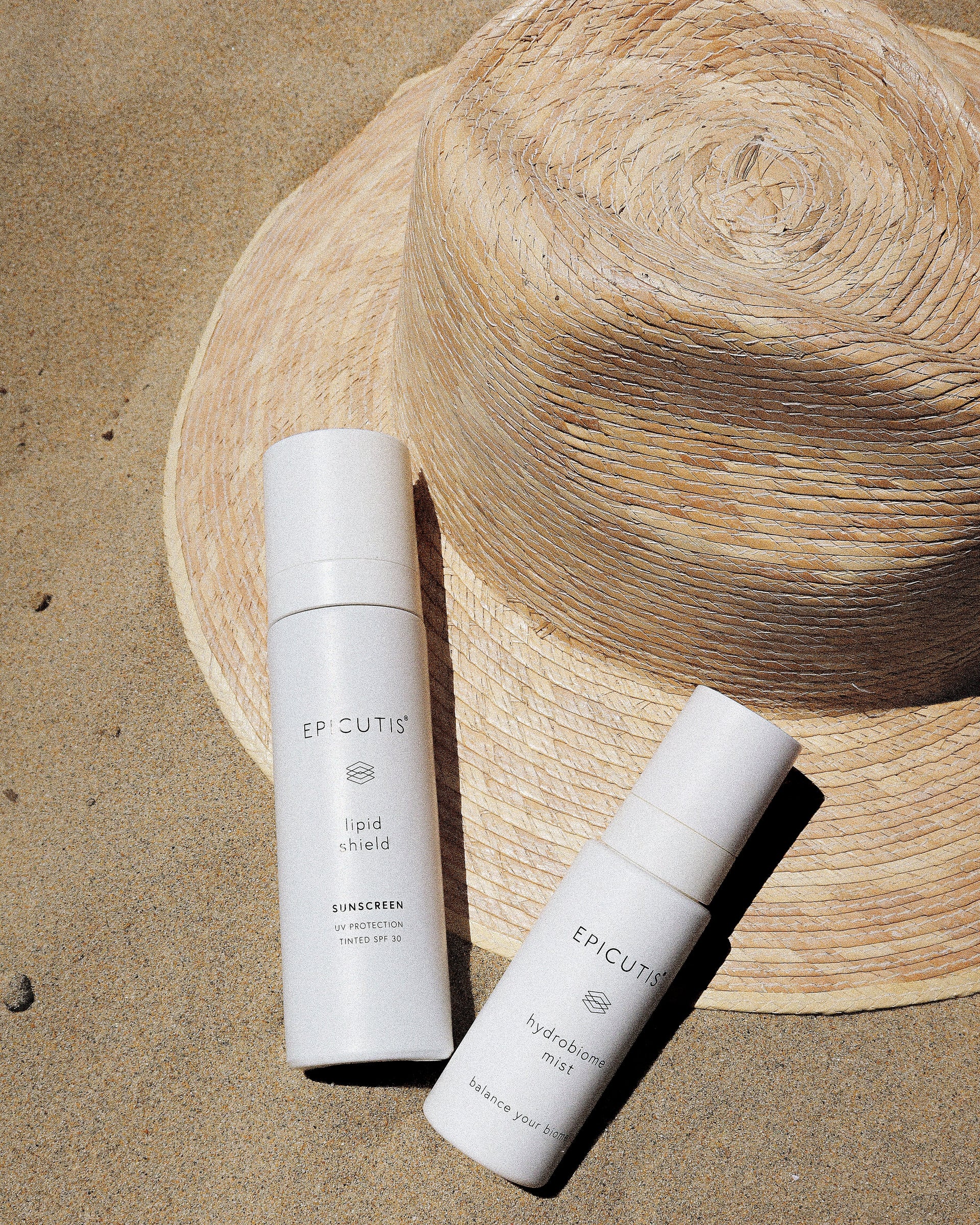Two skincare products on sand with a straw hat in the background
