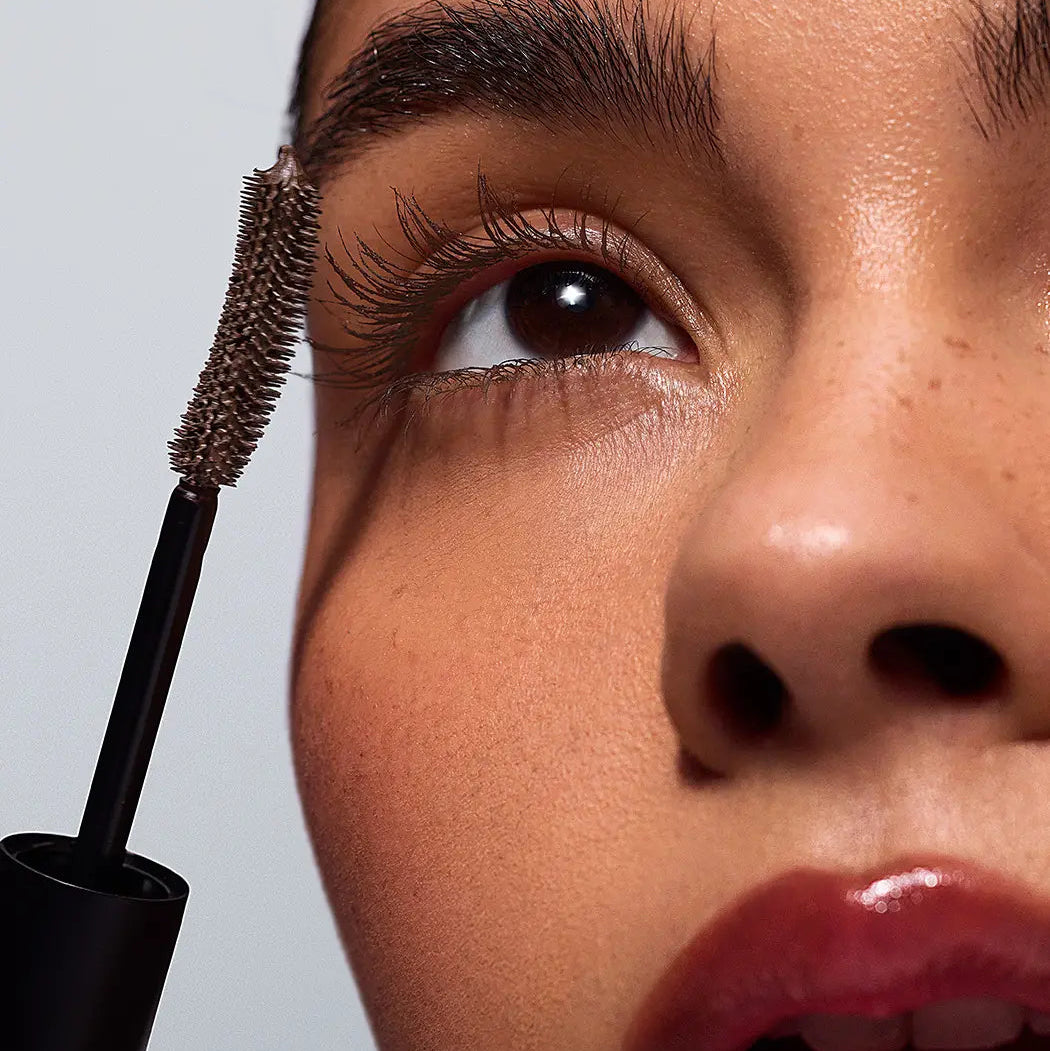 Close-up of a person's face holding a brow mascara wand near the eyebrow, showing the product being applied to dark brows against a light gray background.