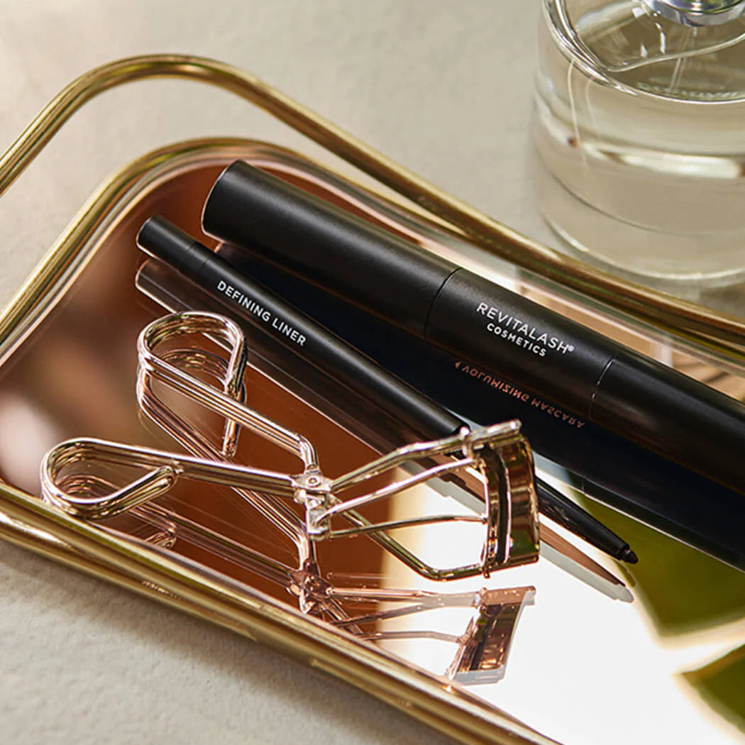 Eyelash curler, eyeliner pencil, and perfume bottle on a gold tray.