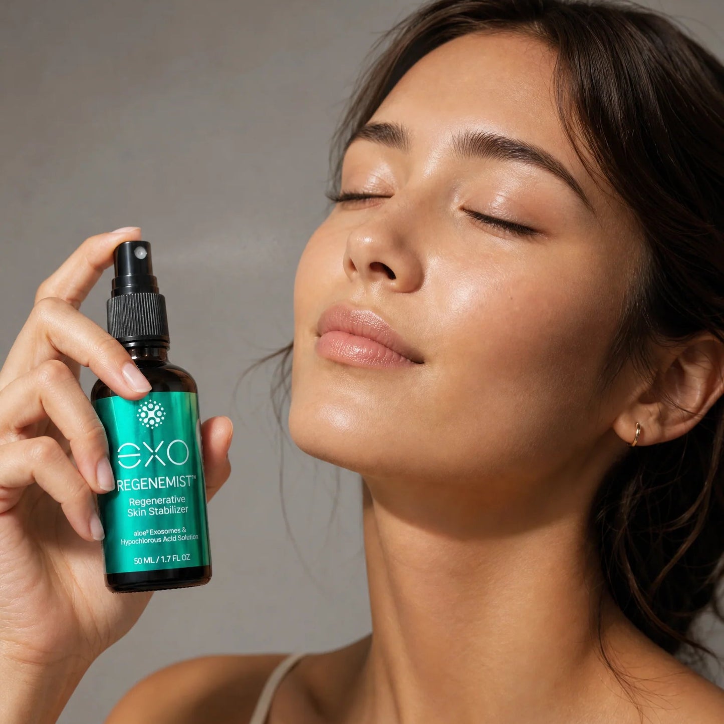 A woman with closed eyes holds a bottle of EXO Regenerist Skin Complex in her hand against a neutral gray background.