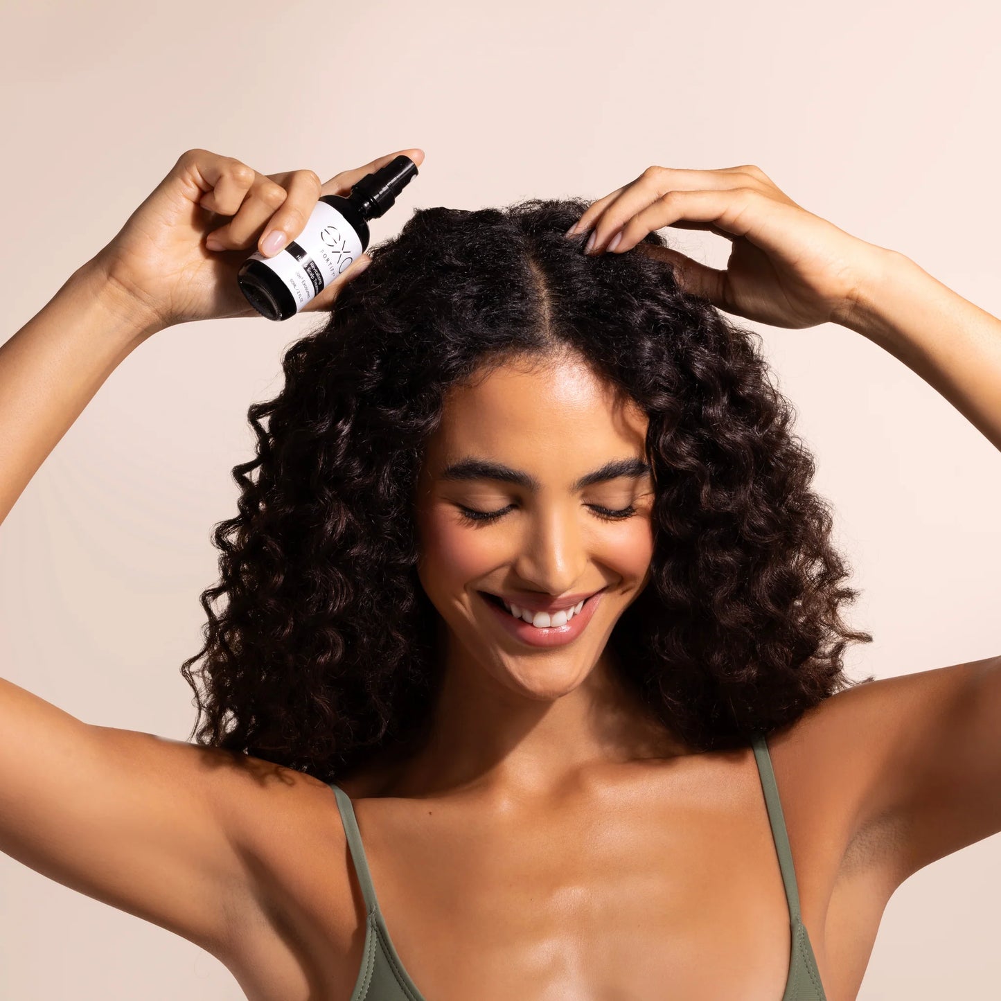 Woman applying hair product to her curly hair with a beige background